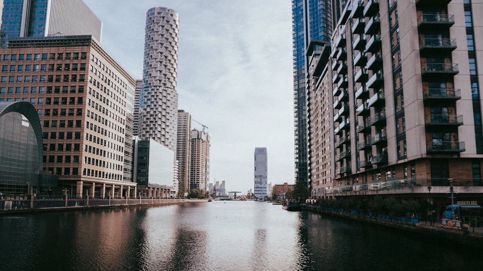 A panoramic view of Canary Wharf Docklands featuring modern high-rise buildings lining a calm waterway. The buildings exhibit a variety of materials, including glass, steel, and concrete, with some structures having reflective surfaces and others with textured facades. The scene is illuminated by natural daylight, creating a mixture of bright and shadowed areas. Visible in the foreground are the water's surface reflecting the surrounding skyscrapers, with some gentle ripples. The background shows a partly cloudy sky. This urban landscape highlights architecture typical of commercial and residential spaces in Canary Wharf, with clean lines and well-maintained exteriors, emphasizing a sense of cleanliness and modernity associated with professional deep cleaning and surface maintenance, as executed by Carpet Cleaners Docklands.