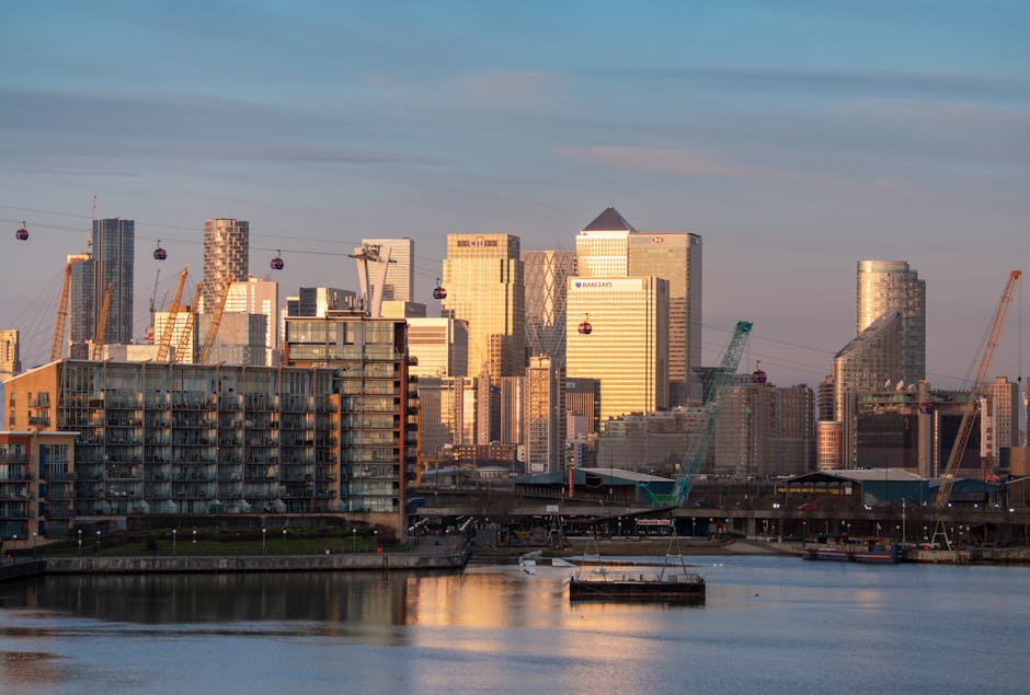 A panoramic view of Canary Wharf in Docklands during sunset, featuring a cluster of modern high-rise office buildings with glass facades reflecting the warm light. Several construction cranes are visible among the skyscrapers, indicating ongoing development. In the foreground, there are mid-rise residential buildings with balconies, situated along the riverbank. The river reflects the cityscape and the soft pinkish sky, with a few small boats moored along the water's edge. The scene captures a mix of commercial and residential architecture, highlighting the vibrant urban atmosphere of Canary Wharf, an area serviced by professional cleaning specialists like Carpet Cleaners Docklands, ensuring cleanliness and hygiene in both residential and business environments.