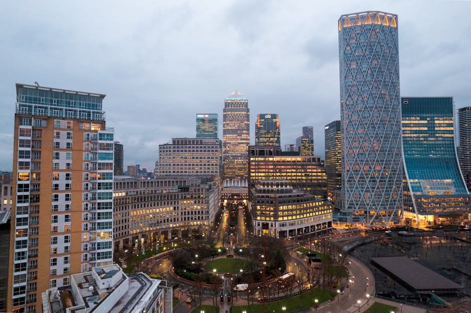 A panoramic view of Canary Wharf Docklands featuring modern high-rise buildings lining a calm waterway. The buildings exhibit a variety of materials, including glass, steel, and concrete, with some structures having reflective surfaces and others with textured facades. The scene is illuminated by natural daylight, creating a mixture of bright and shadowed areas. Visible in the foreground are the water's surface reflecting the surrounding skyscrapers, with some gentle ripples. The background shows a partly cloudy sky. This urban landscape highlights architecture typical of commercial and residential spaces in Canary Wharf, with clean lines and well-maintained exteriors, emphasizing a sense of cleanliness and modernity associated with professional deep cleaning and surface maintenance, as executed by Carpet Cleaners Docklands.