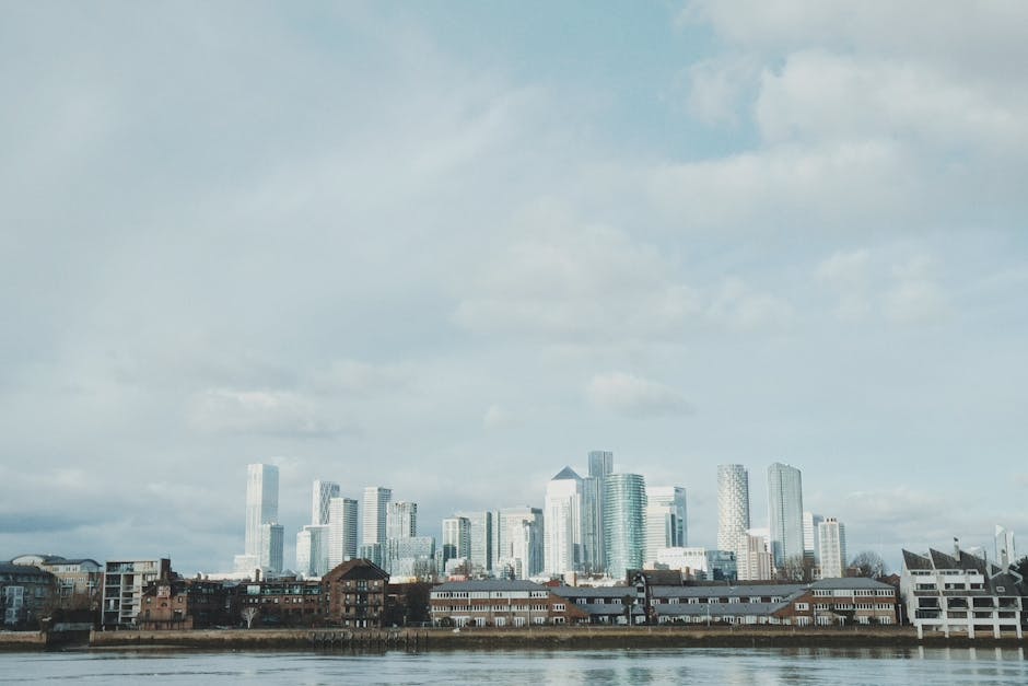 A panoramic view of the Isle of Dogs skyline featuring modern skyscrapers with glass facades reflecting the daylight, set against a backdrop of partly cloudy skies. In the foreground, there are older brick buildings along the riverbank, which appears calm and reflects some of the cityscape. The lighting is natural, highlighting the contrast between the contemporary high-rises and the historical lower structures, emphasizing the area's urban landscape. This image aligns with the expertise of Carpet Cleaners Docklands in surface cleaning and deep cleaning services, showcasing the importance of maintaining a clean and hygienic environment in busy city settings.