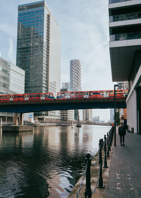 A panoramic view of the Isle of Dogs skyline featuring modern skyscrapers with glass facades reflecting the daylight, set against a backdrop of partly cloudy skies. In the foreground, there are older brick buildings along the riverbank, which appears calm and reflects some of the cityscape. The lighting is natural, highlighting the contrast between the contemporary high-rises and the historical lower structures, emphasizing the area's urban landscape. This image aligns with the expertise of Carpet Cleaners Docklands in surface cleaning and deep cleaning services, showcasing the importance of maintaining a clean and hygienic environment in busy city settings.