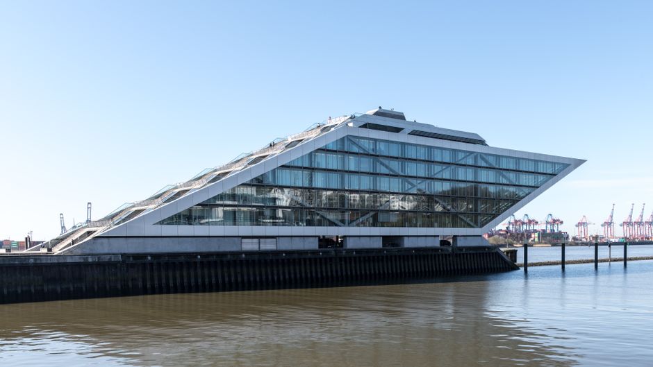 A modern multi-story building with a distinctive slanted design featuring extensive glass windows, located by the water's edge in Docklands. The building's exterior showcases sleek, reflective glass p