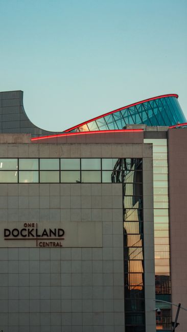 A modern multi-story building with a distinctive slanted design featuring extensive glass windows, located by the water's edge in Docklands. The building's exterior showcases sleek, reflective glass p