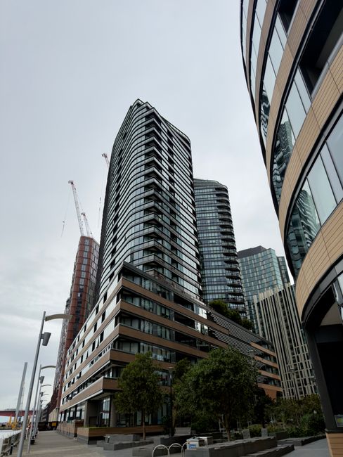 A daytime view of the Docklands area featuring modern high-rise buildings and commercial structures reflecting on the calm river water. The buildings include a mix of glass-fronted offices and residen