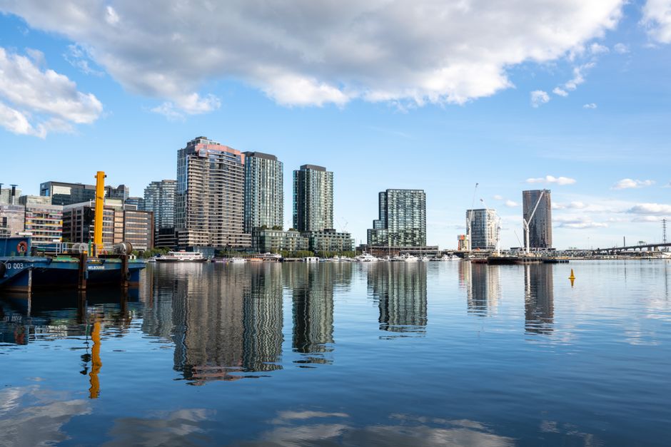 A modern cityscape along a waterfront showing tall, high-rise buildings with glass facades reflecting in the calm water below. Several yachts and boats are docked along the marina, with the sky clear