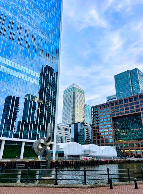 A modern, angular building with large glass windows reflecting the sunset, situated along a calm river in the Docklands area. The building's exterior features metallic and glass surfaces with a sleek,
